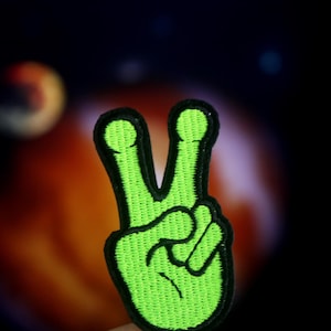 May include: A green peace sign patch with black outlines. The patch is made of fabric and has a textured surface.