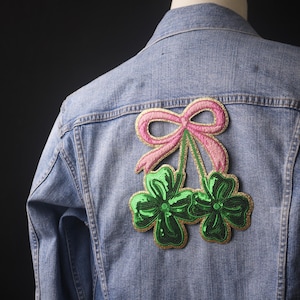 May include: A denim jacket with a green and pink embroidered patch of a clover with a bow on top.