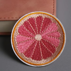 May include: Embroidered patch featuring a grapefruit slice design. The patch has a white border, a light orange outline, and a pink interior with a radial pattern. The patch is approximately 3 inches in diameter.