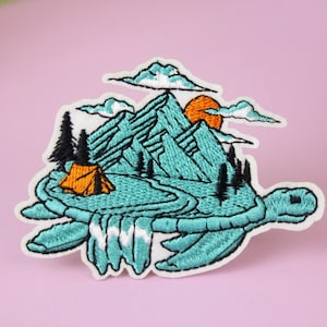 Pode incluir: Um remendo bordado que representa uma tartaruga marinha turquesa carregando uma cena de montanha nas costas. A cena da montanha inclui uma pequena barraca laranja, um caminho sinuoso e um pôr do sol.