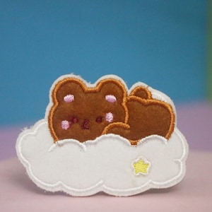 May include: A brown bear with pink cheeks is sleeping on a white cloud with a yellow star. The bear is embroidered on a white fabric.