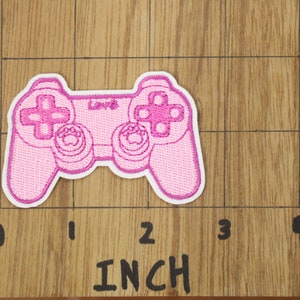 May include: Pink embroidered patch of a video game controller, approximately 5 cm wide.