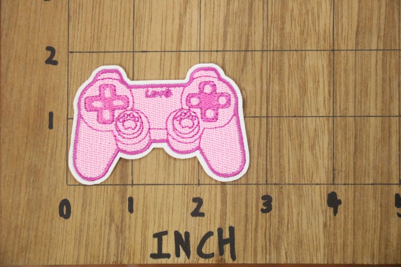 May include: Pink embroidered patch of a video game controller, approximately 5 cm wide.