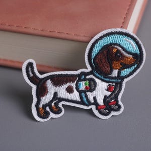 May include: Embroidered patch featuring a dachshund dog in a space suit with a blue helmet. The dog is white with brown spots and a brown head. The patch has a white border and is on a gray surface.