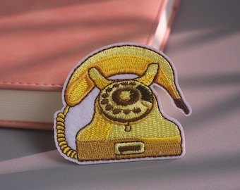 Pode incluir: Um emblema bordado apresentando um telefone de disco amarelo com um mostrador castanho e um fio em espiral. O design é feito com fios em tons de amarelo e castanho sobre um fundo de tecido branco, capturando a forma clássica de um telefone.