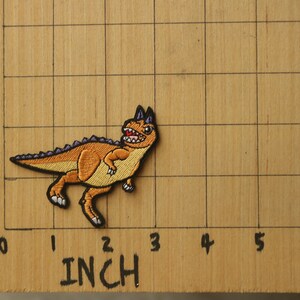 May include: An embroidered dinosaur patch in brown and purple with a toothy grin, measuring approximately 2 inches in length.