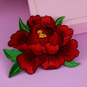 Puede incluir: Parche de flores bordadas de color rojo con hojas verdes. La flor es una peonía con un diseño detallado.