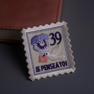 May include: Embroidered patch in the shape of a postage stamp. The design features a purple pansy, a white dove carrying a letter, and the number 39. The text "JE PENSE À TOI" is also included.