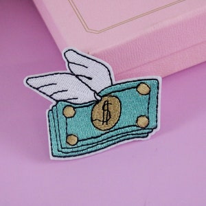 Puede incluir: Un parche bordado para planchar de un montón de billetes con alas. Los billetes son verdes con un signo de dólar dorado en el centro. Las alas son blancas.