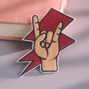 Può includere: Una toppa ricamata raffigurante una mano color marrone chiaro che fa il segno del rock and roll, sovrapposta a una forma di fulmine rosso frastagliato con un bordo nero. Il design è realizzato con ricami dettagliati, contorni netti e superficie piatta.