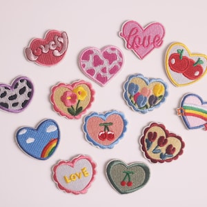 Può includere: Una collezione di toppe ricamate a forma di cuore in vari colori e design. Alcuni cuori presentano motivi floreali, ciliegie, arcobaleni e la parola "love". Altri hanno stampa di mucca o la parola "lovely".