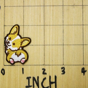 Może przedstawiać: Naszywka haftowana przedstawiająca brązowo-białego corgiego siedzącego na tylnych łapach z wystawionym językiem. Naszywka ma około 2,5 cm wysokości i 2,5 cm szerokości.