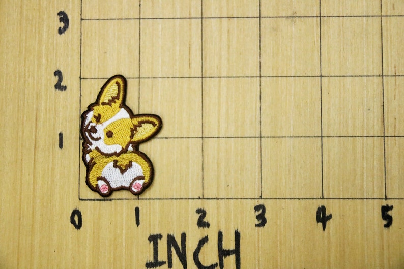 Może przedstawiać: Naszywka haftowana przedstawiająca brązowo-białego corgiego siedzącego na tylnych łapach z wystawionym językiem. Naszywka ma około 2,5 cm wysokości i 2,5 cm szerokości.