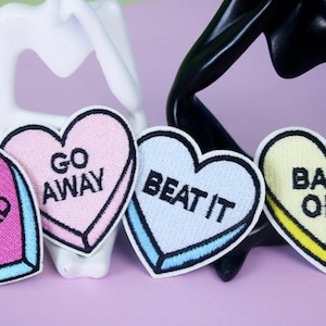 Peut inclure: Quatre patchs thermocollants en forme de cœur avec des dictons différents. Les patchs sont rose, bleu clair, jaune et violet. Les dictons sont "Drop Dead", "Go Away", "Beat It", et "Back Off".