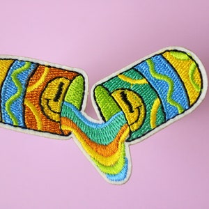 Könnte beinhalten: Gestickter Aufnäher mit zwei pillenförmigen Behältern mit Smiley-Gesichtern, aus denen ein Regenbogen aus Farben quillt. Die Behälter haben gelbe, blaue und grüne Streifenmuster. Der Aufnäher hat einen weißen Rand auf rosa Hintergrund.