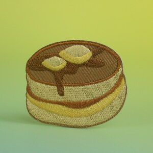 May include: A brown embroidered patch of a stack of pancakes with a brown syrup drizzle and yellow butter on top.