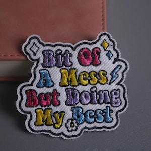 Puede incluir: Parche bordado con el texto "Bit Of A Mess But Doing My Best" en letras coloridas estilo burbuja. El parche tiene un borde blanco y está decorado con estrellas, un rayo y una flor.
