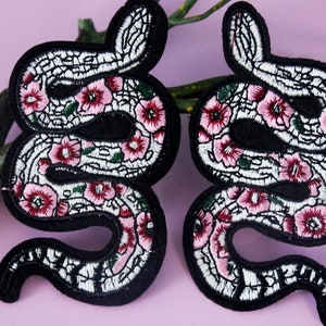 May include: Two black embroidered iron-on patches of snakes with pink flowers. The snakes are curled up and have a white and green pattern.