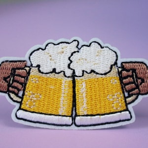 May include: Embroidered patch of two beer mugs clinking together, with foam on top, held by two brown hands. The mugs are yellow with white outlines and the foam is white.