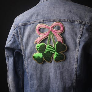 Puede incluir: Una chaqueta vaquera con un parche bordado de tres tréboles en verde y dorado, coronado con un lazo rosa.
