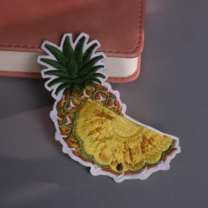 May include: Embroidered pineapple patch with green leaves and a yellow fruit slice. The patch has a white border and is placed on a gray surface next to a pink notebook.