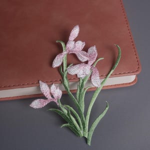 May include: Embroidered floral patch featuring a cluster of pink and white irises with green stems and leaves. The patch is placed on a closed brown leather-bound notebook, set against a gray background.