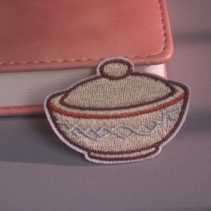Può includere: Una toppa ricamata raffigurante una ciotola beige con coperchio, un bordo marrone scuro e un motivo intrecciato grigio chiaro sulla base. Il design è delineato con filo marrone scuro e presenta un bordo bianco con cuciture dettagliate.