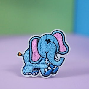 Pode incluir: Um remendo bordado de um elefante azul com orelhas cor-de-rosa. O elefante está andando e tem uma pequena cauda laranja.