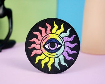 Op de afbeelding: Een ronde zwarte patch met een geborduurd ontwerp van een zon met een groot oog in het midden. De zon is gemaakt van roze, gele, groene en paarse stralen.