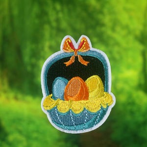 May include: A blue and white embroidered patch of a basket with a yellow bow and three colorful eggs inside.