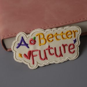 Pode incluir: Um remendo branco bordado com o texto "A Better Future" em fio colorido. O remendo tem a forma de uma nuvem e é perfeito para adicionar um toque de fantasia a roupas, bolsas ou outros acessórios.