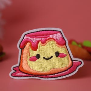 May include: A pink and yellow embroidered iron-on patch of a cartoon pudding with a happy face. The pudding has a pink swirl on top and a white outline.