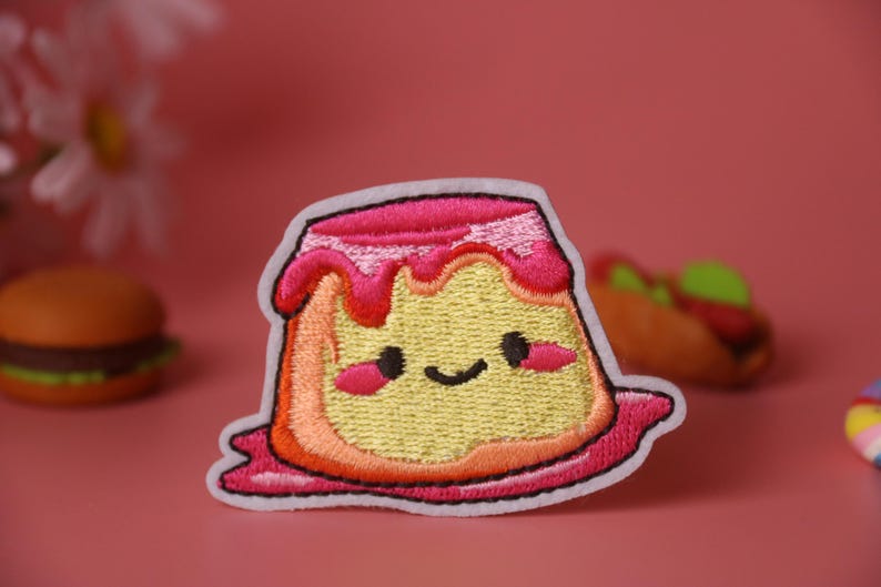 May include: A pink and yellow embroidered iron-on patch of a cartoon pudding with a happy face. The pudding has a pink swirl on top and a white outline.