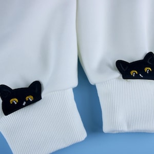 Puede incluir: Sudadera blanca con bordado de cara de gato negro en los puños. Las caras de gato tienen ojos amarillos.
