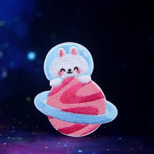 May include: Embroidered iron-on patch featuring a white bunny wearing a blue astronaut suit and holding a pink planet with a blue ring around it.