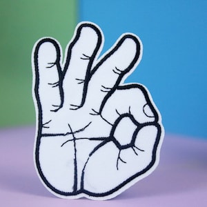 May include: A white embroidered patch of a hand making the OK sign. The hand is outlined in black thread.