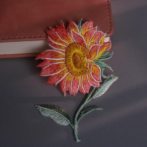 May include: Embroidered sunflower patch with orange, yellow, and white petals, green stem and leaves. The patch is placed on a grey surface next to a pink leather-bound notebook.