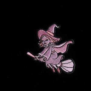 Puede incluir: Parche bordado con una bruja montada en una escoba. La bruja está representada en tonos rosas y morados, con un sombrero puntiagudo y una capa. El parche está sobre un fondo negro.