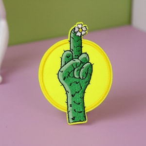 Könnte beinhalten: Ein grüner Kaktus in Form eines Mittelfingers mit einer weißen Blüte oben. Der Kaktus befindet sich auf einem gelben Kreisfleck.