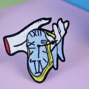 Puede incluir: Un parche bordado para planchar que presenta una mano sosteniendo un reloj derritiéndose. El reloj es azul con números romanos negros y detalles amarillos. La mano es blanca con detalles rojos.