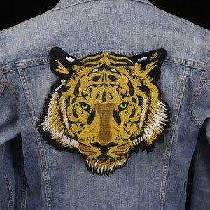 Op de afbeelding: Een close-up van een geborduurde tijgerkop patch in geel en zwart. De patch is genaaid op een blauw denim jack.
