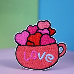 May include: An embroidered red mug with the word "LOVE" on the front, filled with pink and red hearts.