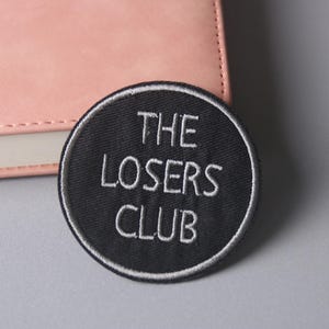 Op de afbeelding: Een geborduurde zwarte en witte patch met de tekst "THE LOSERS CLUB" in witte letters op een zwarte achtergrond.