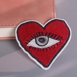 Pode incluir: Patch bordado em forma de coração vermelho com borda branca. O coração apresenta um design detalhado de olho no centro, com íris cinza e cílios brancos. O patch está colocado sobre uma superfície cinza, ao lado de um caderno rosa.