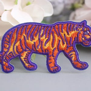 Op de afbeelding: Een geborduurde strijk-op patch van een oranje en paarse tijger. De tijger loopt naar rechts en heeft een gedetailleerd gestreept patroon.