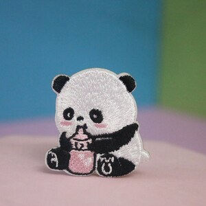 May include: A white and black embroidered panda bear patch with pink accents. The panda is holding a pink bottle in its paws.