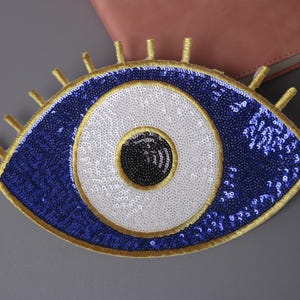 Puede incluir: Un amuleto de ojo turco azul y blanco con ribetes dorados y lentejuelas. El amuleto es un símbolo popular de protección contra la energía negativa.