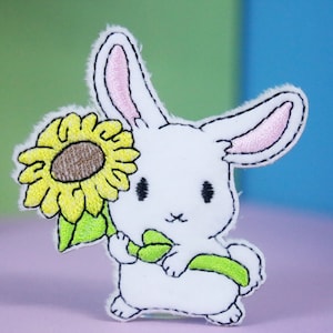 May include: A white bunny patch with a sunflower. The bunny has pink inner ears and holds a yellow sunflower with a brown center and green leaves. The patch is embroidered with black thread.