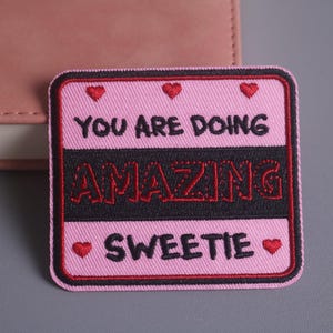 Peut inclure: Écusson rose brodé avec les mots "YOU ARE DOING AMAZING SWEETIE" en noir et rouge. L'écusson a une bordure noire et de petits cœurs rouges. L'écusson est rectangulaire.