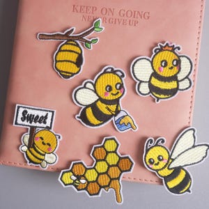 May include: Embroidered bee patches in yellow, black, and white, including a honeycomb, a beehive, and bees with crowns and holding honey. The patches are on a pink surface with the text "KEEP ON GOING NEVER GIVE UP".